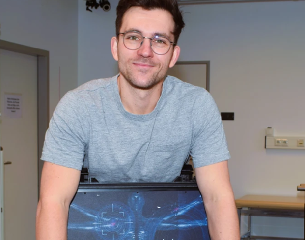 A man in a gray t-shirt and glasses stands, smiling, behind a computer displaying an X-ray image in an office setting.