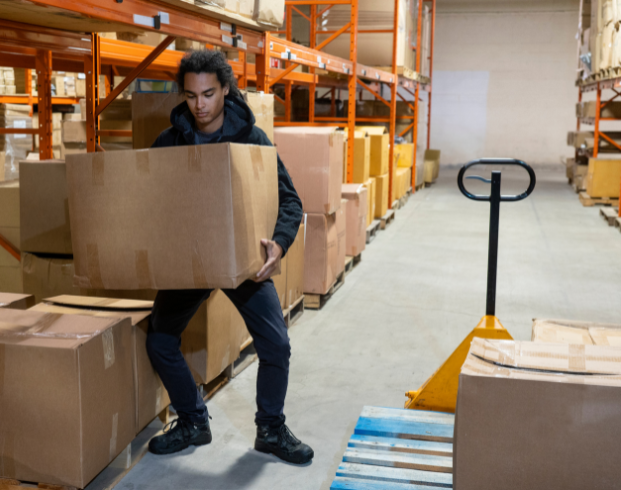 Person lifting a large cardboard box in a warehouse, surrounded by shelves filled with additional boxes. A pallet jack is visible nearby on the floor.