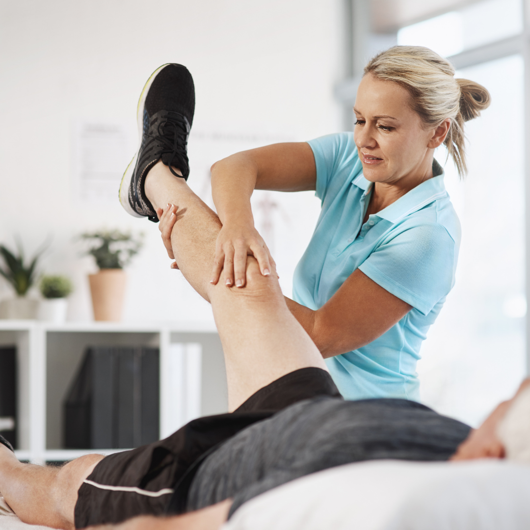 A physical therapist in a blue shirt assists a person lying on a table by lifting and supporting the individual's leg.