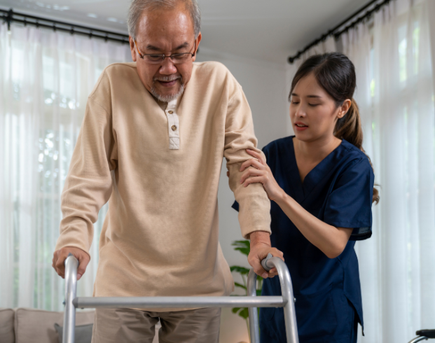 An elderly man uses a walker while a young caregiver in blue scrubs assists him inside a well-lit room with white curtains.