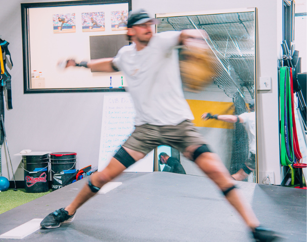 A person in sportswear is captured mid-throw in an indoor training space. The motion blur emphasizes the action. Behind them are sports equipment, a whiteboard, and a mirror reflecting the scene.