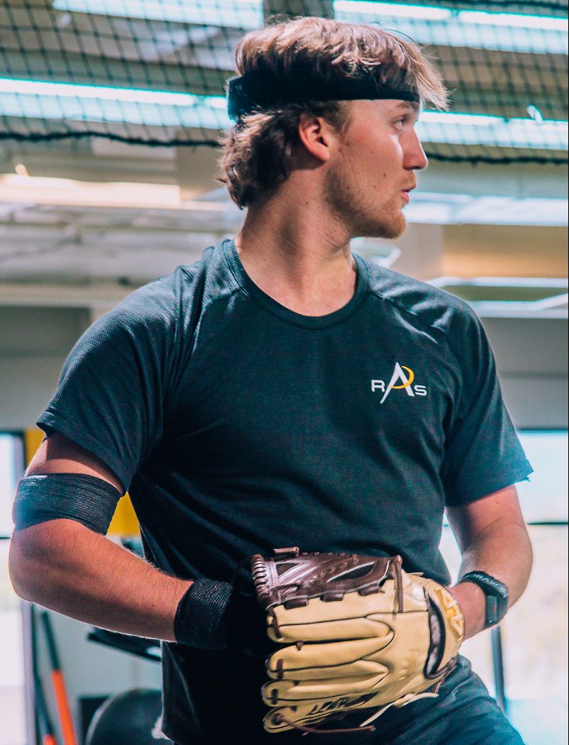 A baseball player wearing a dark t-shirt, with a headband and arm wrap, holding a glove, standing indoors with netting and lights visible in the background.