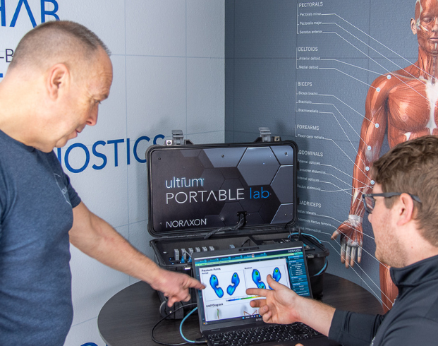 Two men in conversation analyze foot pressure data on a laptop screen, with an Ultium Portable Lab system nearby and an anatomical chart of muscles in the background.