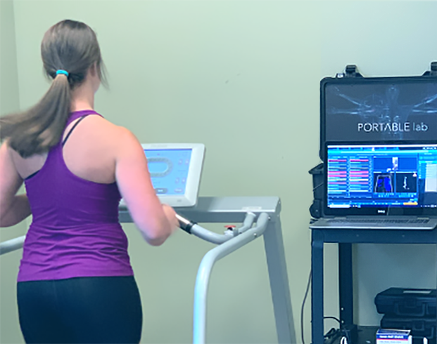 A person with a ponytail is running on a treadmill, wearing a purple tank top. A laptop with the screen displaying the text "PORTABLE lab" is on a small shelf next to the treadmill.