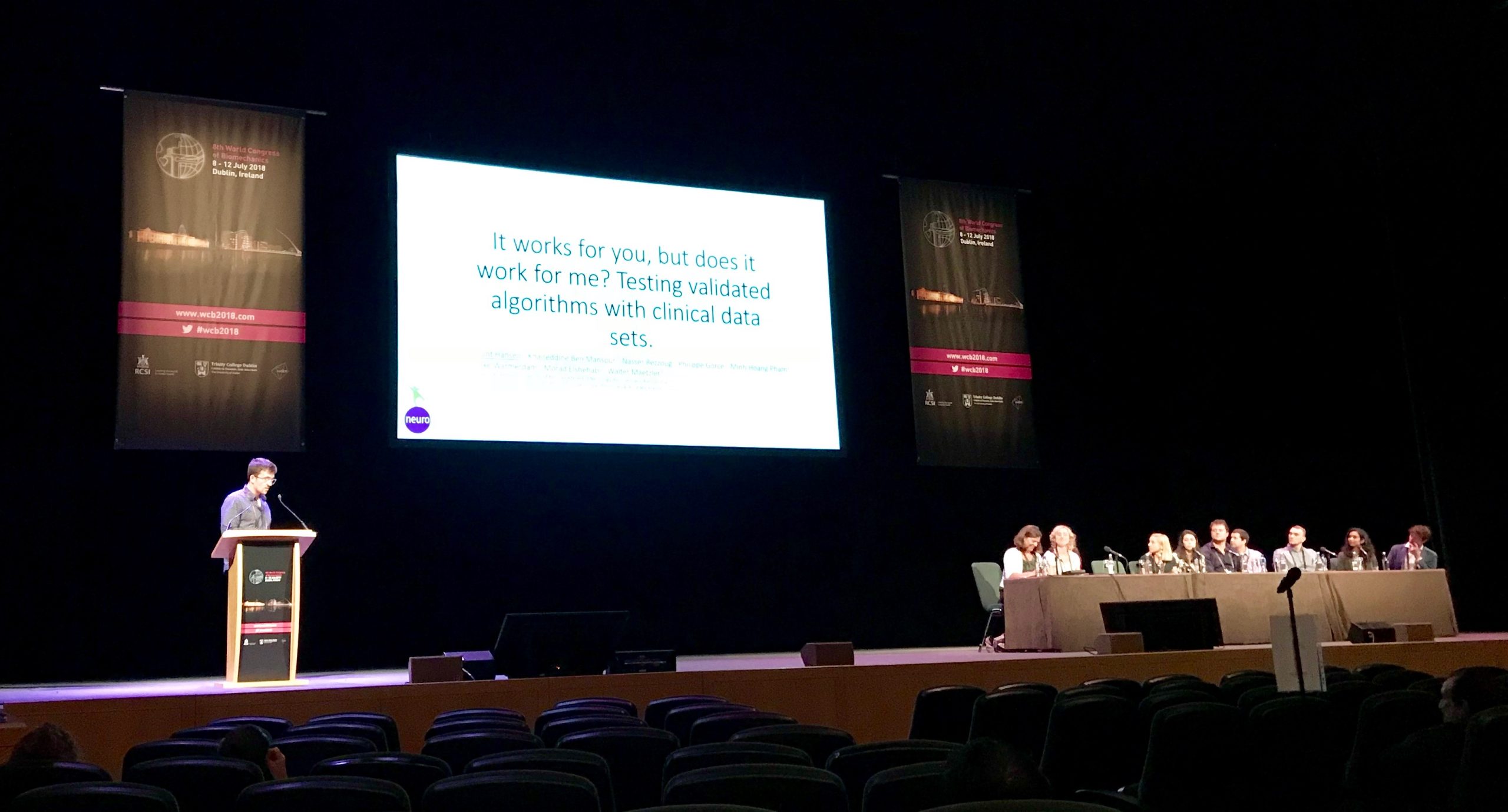 A speaker presents at a podium on stage with a projected slide that reads, "It works for you, but does it work for me? Testing validated algorithms with clinical data sets." A panel of people sits at a table.