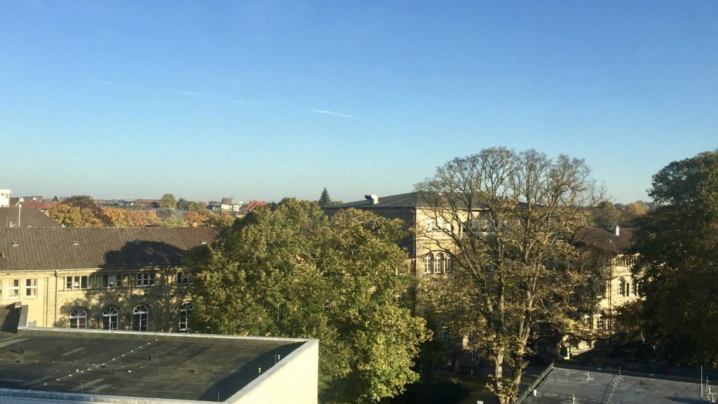 View of several large buildings surrounded by trees under a clear blue sky on a sunny day.