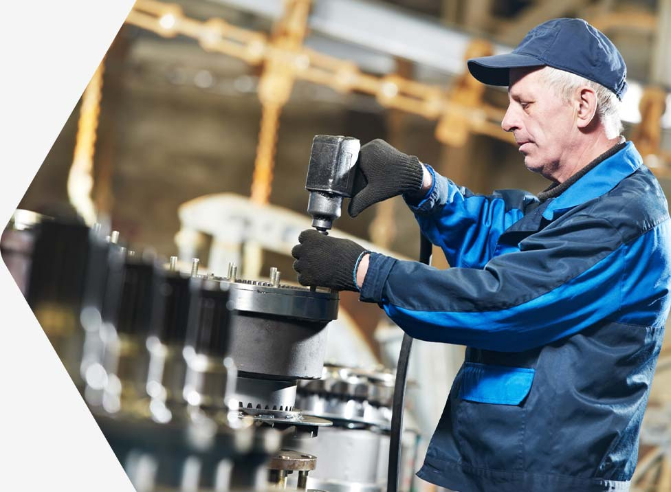 An older man wearing a blue cap and work uniform operates a power tool in an industrial setting.