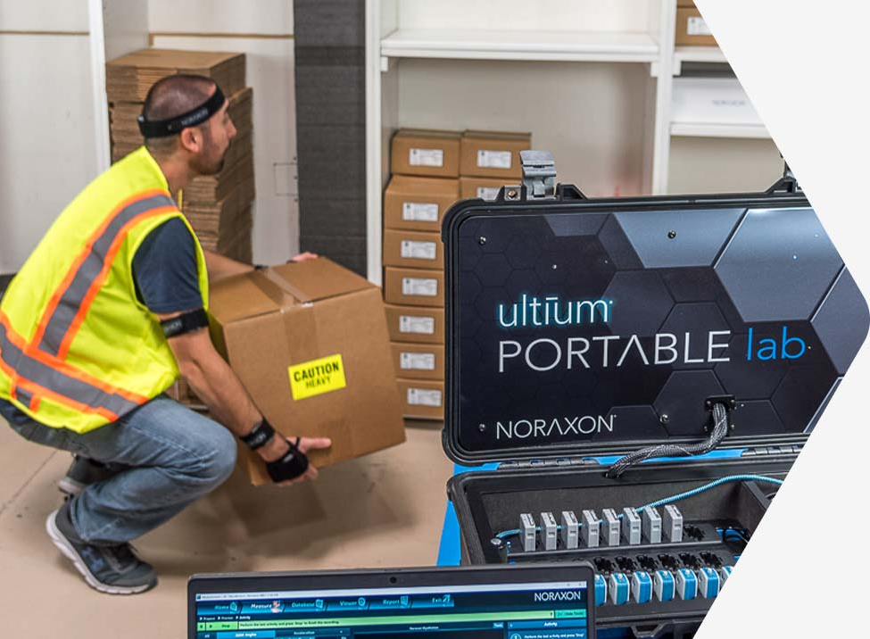 A worker in a high-visibility vest lifts a box labeled "Caution Heavy" next to a NORAXON Ultium Portable Lab equipment setup in an indoor setting.