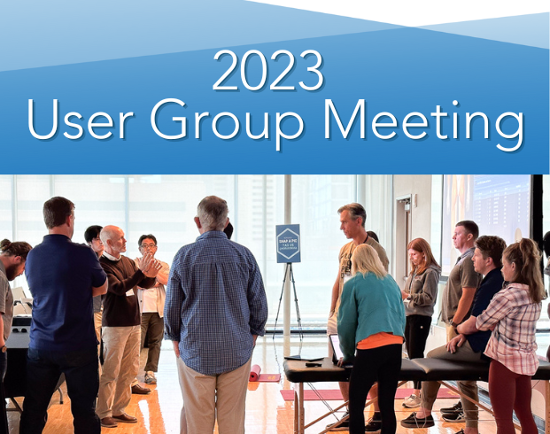 A group of people stand in a semi-circle, engaging in discussion during the 2023 User Group Meeting in a well-lit room.