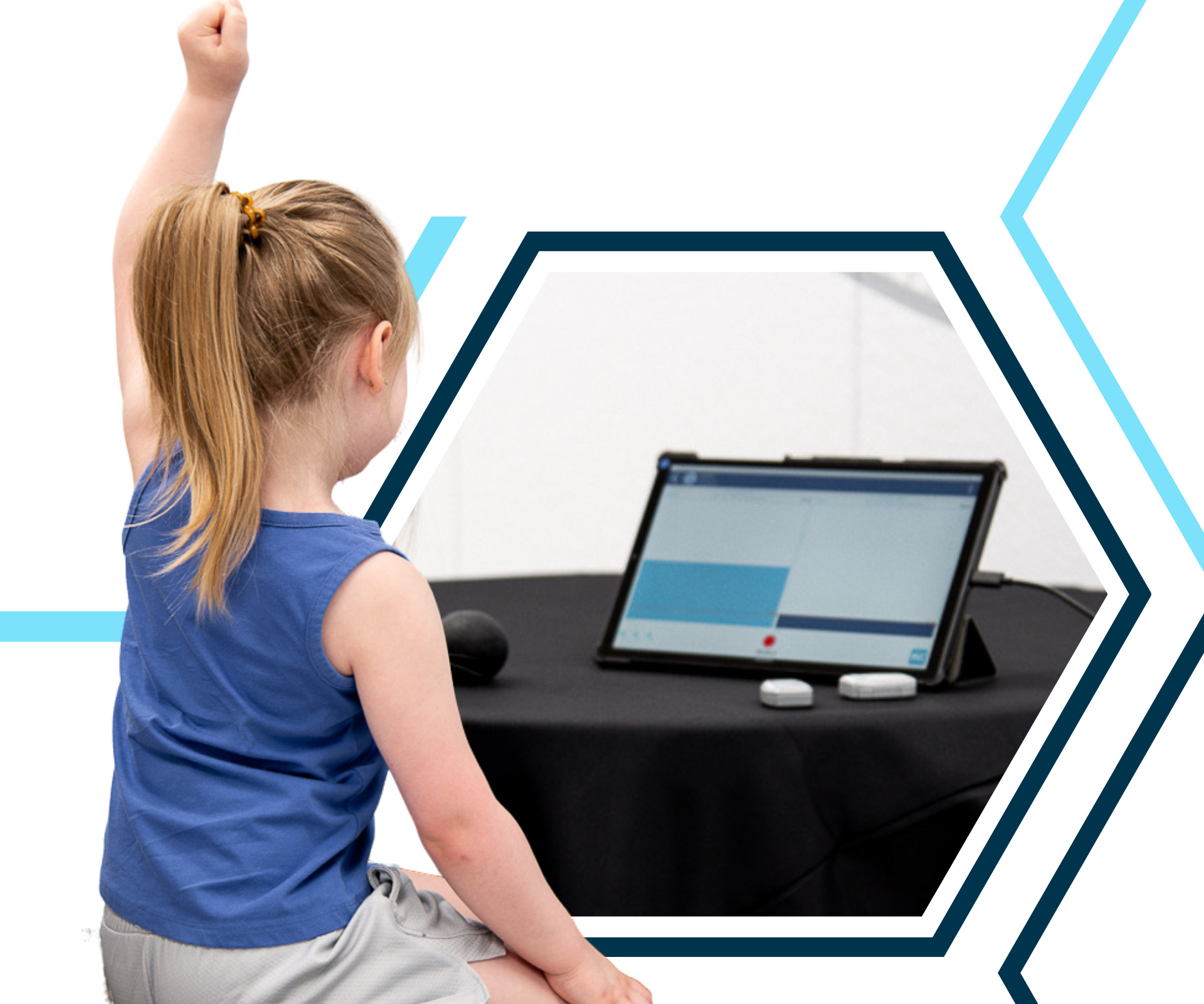 A young girl with a ponytail raises her hand while looking at a tablet on a table.