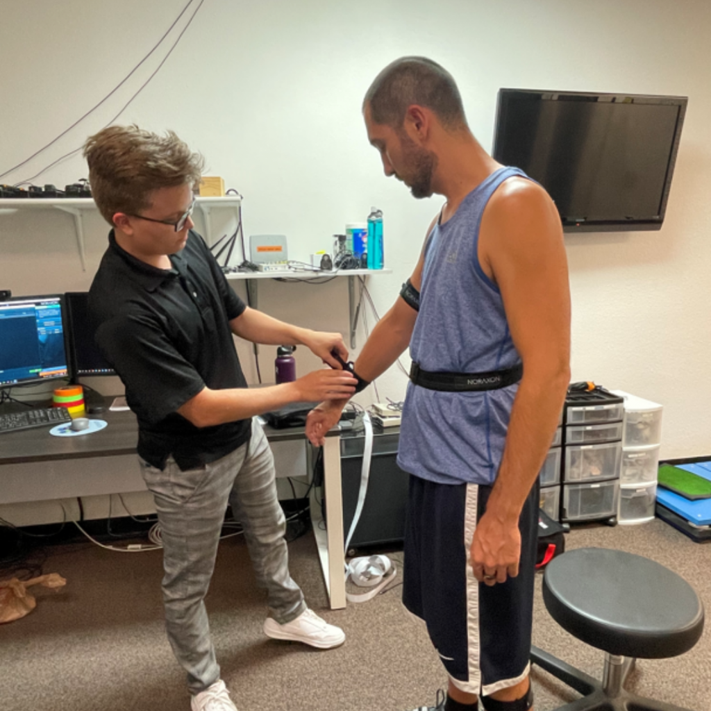 A technician adjusts sensors and straps on a man's arm and torso in a room with a desk, computer, monitor, and cabinets.