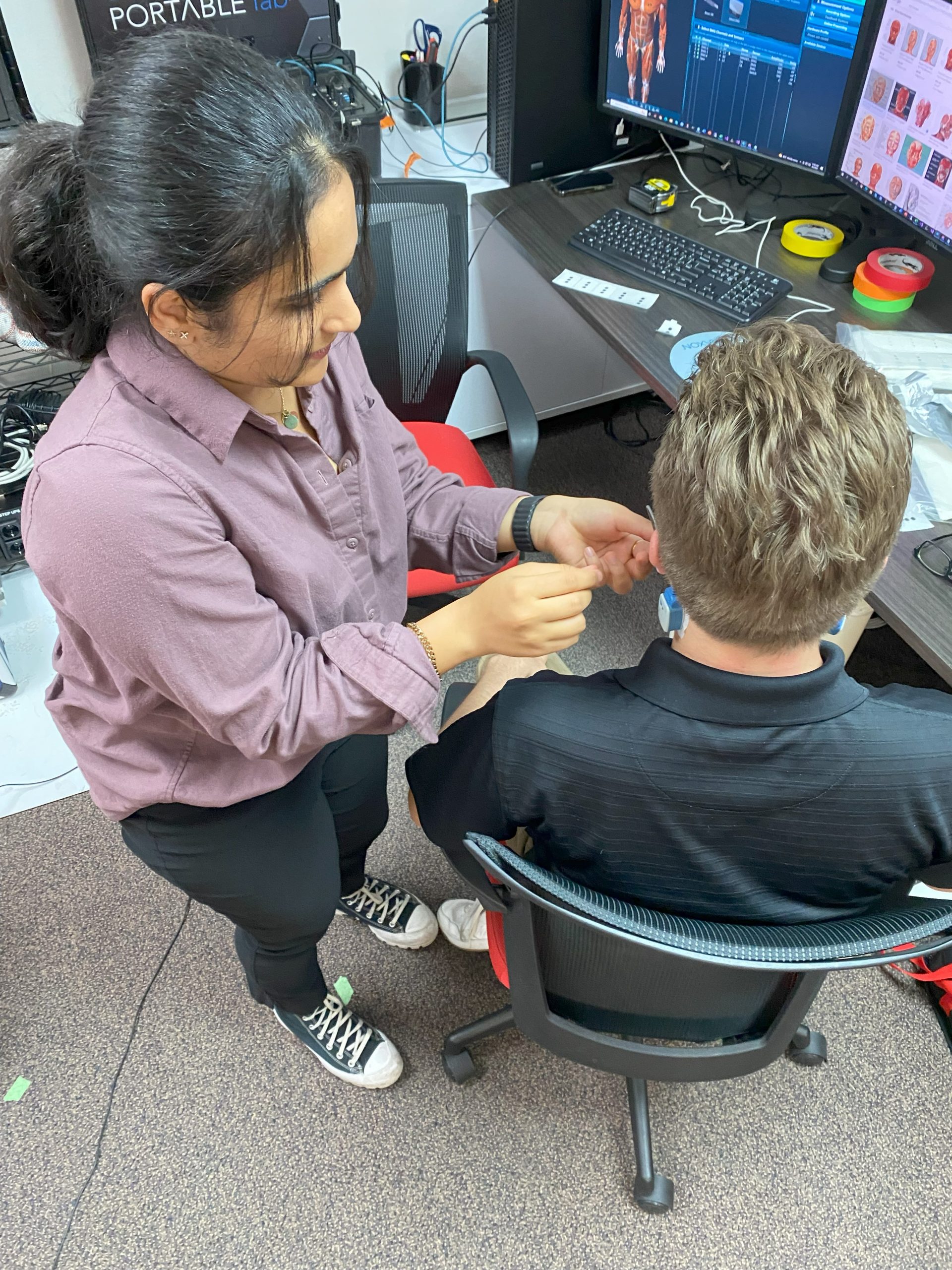 A woman stands next to a seated man, attaching a small device to his neck with wires connected to a nearby computer screen displaying data. The setting is an office with various electronic equipment.