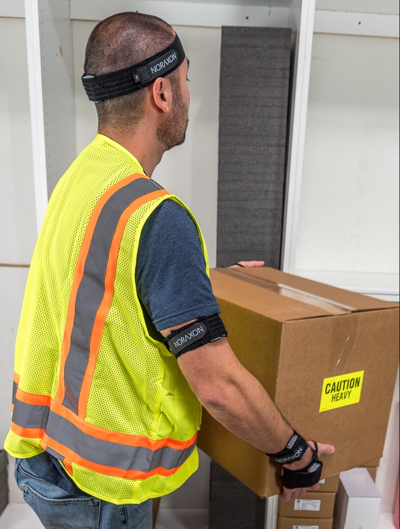 A person wearing a high-visibility vest, headband, and wristbands labeled with "NORAXON" is lifting a brown box with a "CAUTION HEAVY" label.