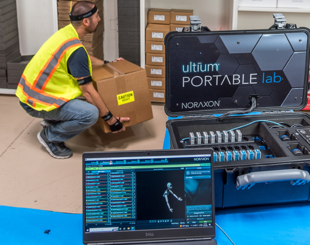 A person in a high-visibility vest lifts a box labeled "Caution" in a workspace. In the foreground, the Noraxon Ultium Portable Lab and a laptop are set up for motion analysis.