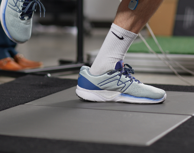 Close-up of a person wearing white and blue athletic shoes, stepping on a gray platform with a sensor attached to their calf.