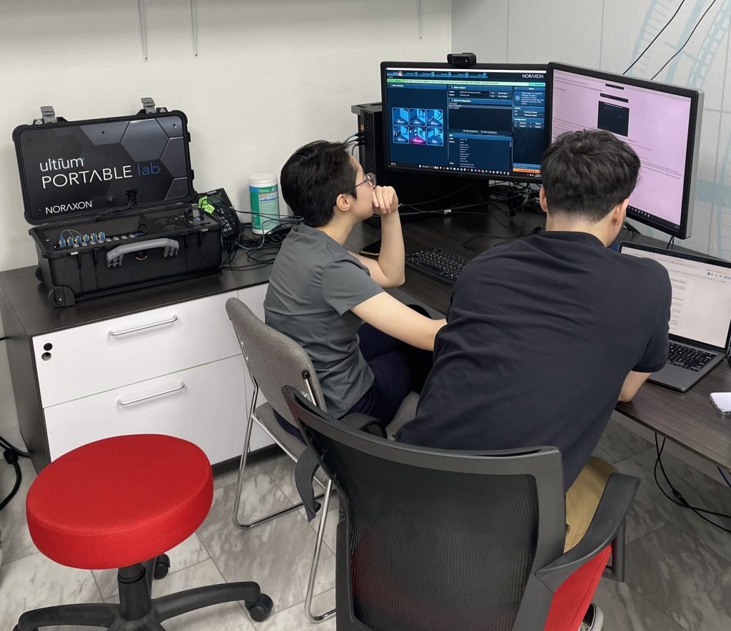 Two individuals sit at a desk in front of computer monitors displaying technical information. A portable lab kit labeled "ultium PORTABLE lab Norton" is on a small table beside them.