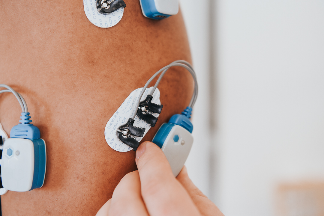 Close-up of a person's skin with several blue and white electrode pads attached, connected by grey wires, with a hand adjusting one of the pads.