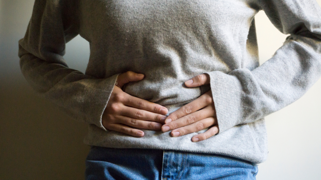 A person wearing a gray sweater and blue jeans holds their stomach with both hands, suggesting they may be experiencing abdominal discomfort.