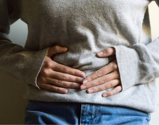 A person wearing a gray sweater and blue jeans is holding their abdomen with both hands.