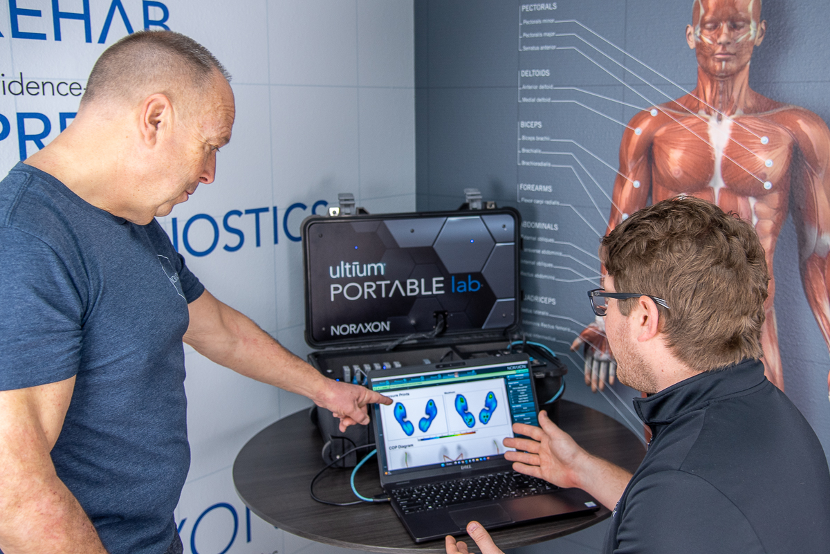 Two men analyze foot pressure data on a laptop connected to a device labeled "ultiuM PORTABLE lab" with an anatomical chart in the background.