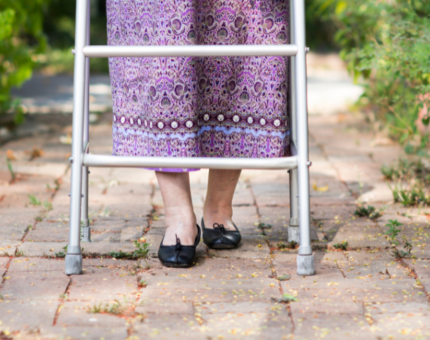 A person using a walker stands on a brick pathway, wearing black shoes and a purple patterned garment.