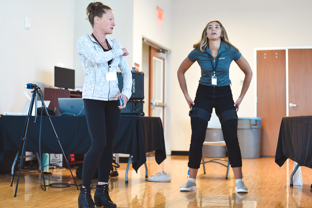 Two women are standing in a room with computer equipment. One is speaking and gesturing, and the other is wearing a powered exoskeleton and appearing to adjust its fit.