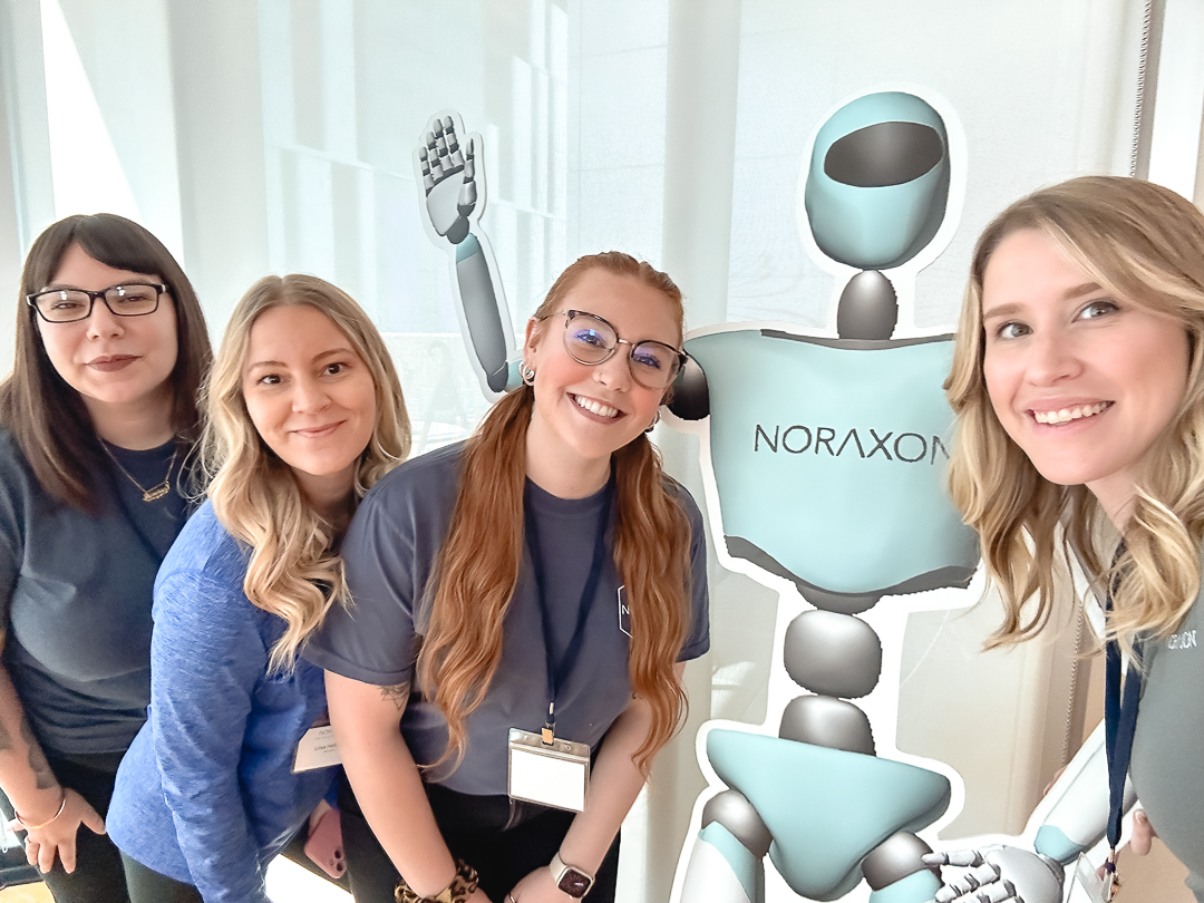 Four women pose next to a robot illustration, with one of them taking a selfie. The robot has "NORAXON" written on its chest.