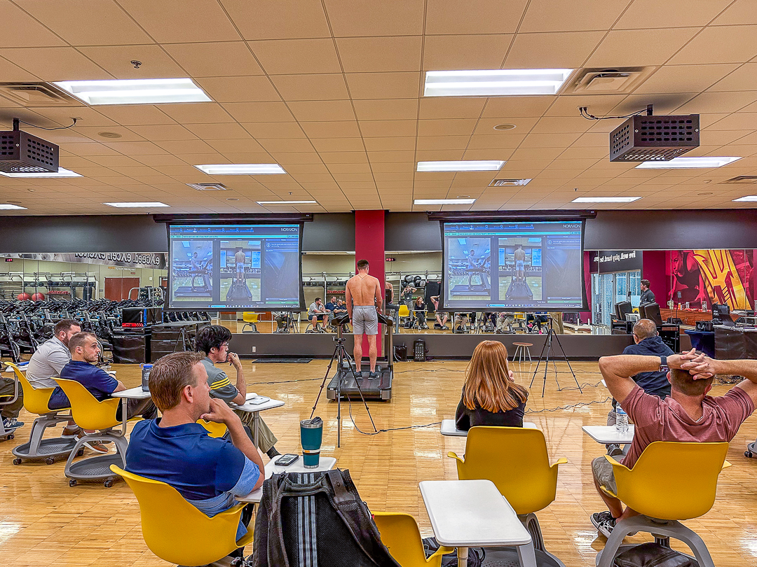 A group of people seated in a gym classroom watch a man stand shirtless on a platform in front of two large projection screens displaying mirrored fitness data.