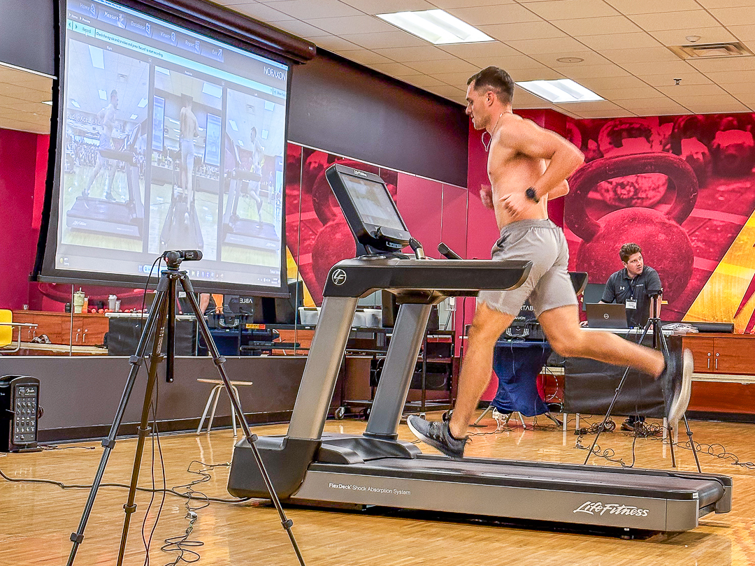 A man runs on a treadmill in a gym, with video capture equipment and a large screen showing his movements in the background. Another person is seated at a desk with a laptop.