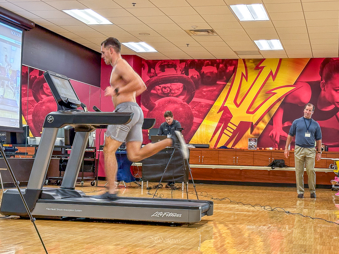 A man runs on a treadmill in a gym setting, observed by two other individuals. The background features a large graphic with athletic imagery.