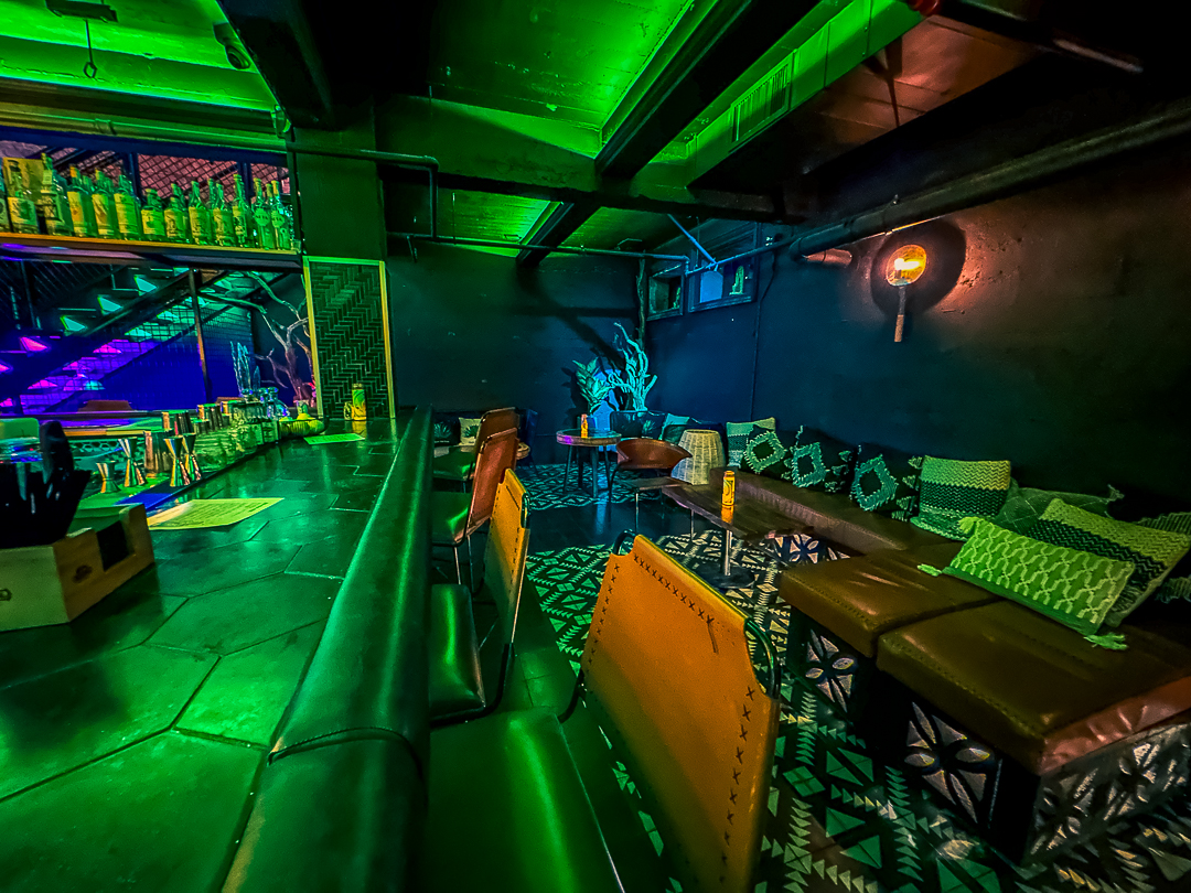 Dimly lit lounge with green and blue lighting, featuring cushioned seating and tables, a long bar counter on the left, and colorful decor.