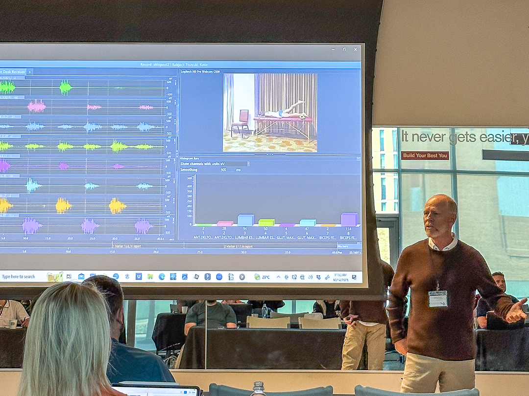 A man stands in front of a large screen displaying colorful waveform data and an image. He appears to be giving a presentation to an audience in a room with glass walls.