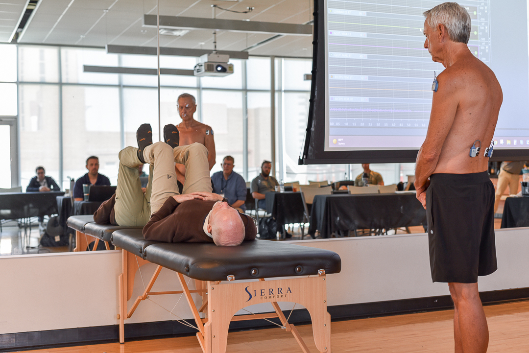 A shirtless man and a man lying on a table with electrodes participate in a study while seated individuals observe from the background.