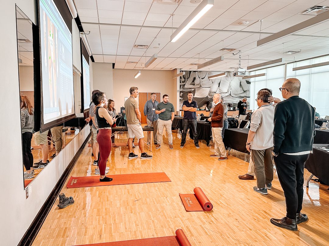 A group of people stand in a circle in a room with yoga mats and a large screen, engaged in conversation or instruction. The room has mirrors, wooden flooring, and tables in the background.