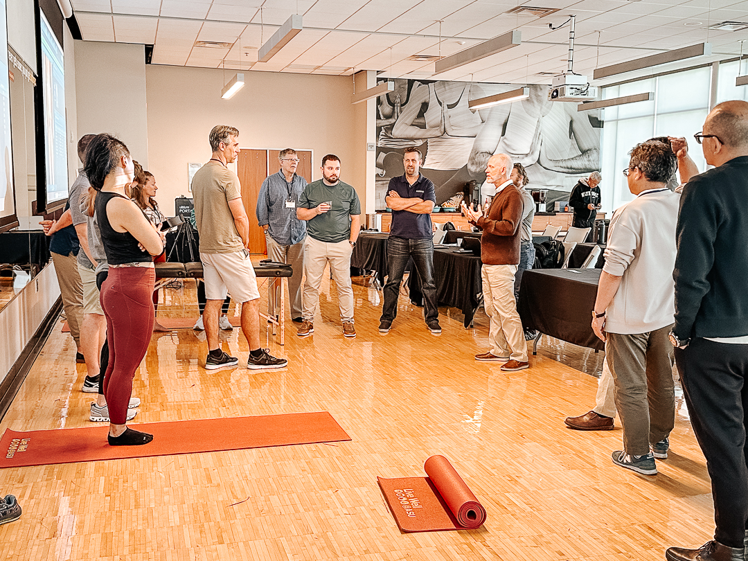 A group of people stands in a circle in a bright room with wooden floors, engaging in a discussion. Two yoga mats are on the floor. A presentation screen is visible in the background.