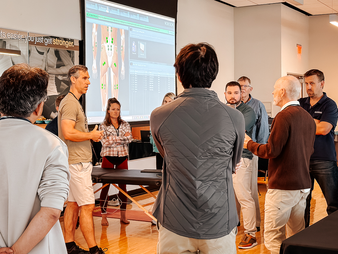 A group of people, some standing and some sitting, listen to a man presenting near a large screen displaying anatomical imagery in a meeting room.