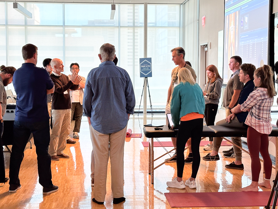 A group of people are gathered around a presentation in a brightly lit room. Some are standing, while others are using a massage table as a prop. A screen displaying graphics is visible in the background.