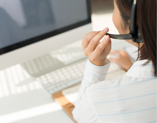 Person wearing a headset, holding the microphone, and working on a desktop computer in a bright office setting.