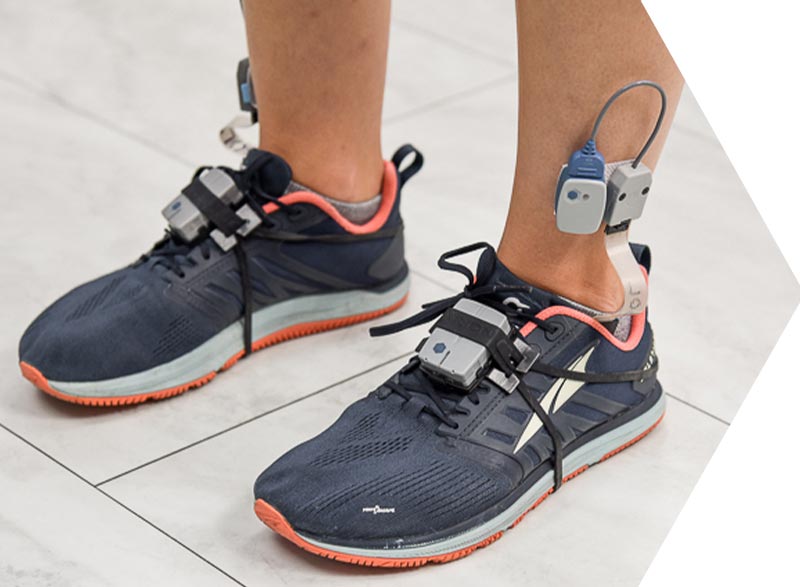 A person wearing dark athletic shoes, equipped with electronic devices and pressure insoles attached to the ankles and shoelaces, stands on a light-colored floor.